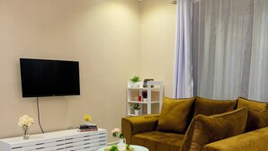 A living room with a brown sofa, a white side table, a flat-screen TV, a white storage unit, and a window with curtains. - Luxury One Bedroom Apartment Kampala Uganda. (Kampala)