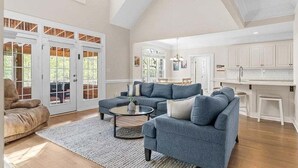 A spacious living area with a high ceiling, a kitchen with white cabinets, and a dining area with a table and chairs.