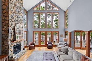 A spacious living room with a stone fireplace, large windows, and a high ceiling.