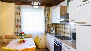 A compact kitchen with a stove, oven, and modern cabinetry. A dining table with yellow chairs and a vase of flowers is positioned near a window with sheer curtains. - Bungalow on Lake Klopein With Garden (Sankt Kanzian am Klopeiner See)