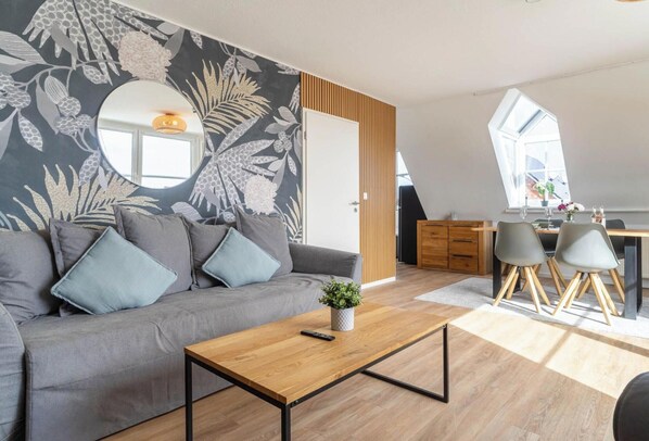 A modern living room with a grey sofa, a wooden coffee table, and a dining area with a table and chairs. - Ferienwohnung in St.peter-ording (St. Peter-Ording)