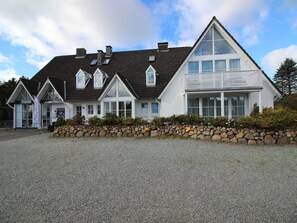 A two-story building with a stone wall in front, a gravel driveway, and a clear sky. - Ferienwohnung in St.peter-ording (St. Peter-Ording)
