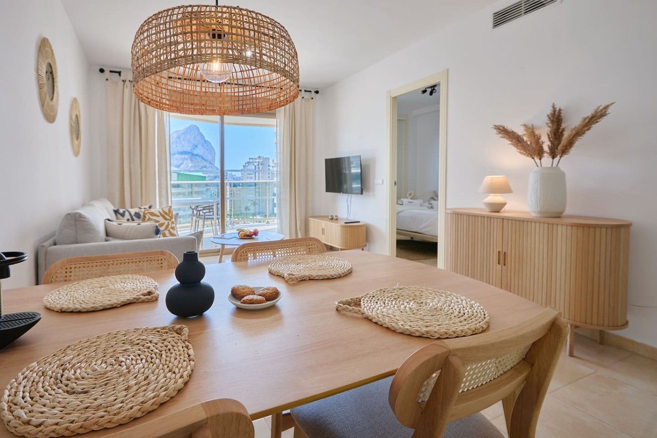 Una sala de estar moderna con comedor, que presenta una gran mesa de madera, individuales tejidos y una lámpara de techo de ratán.