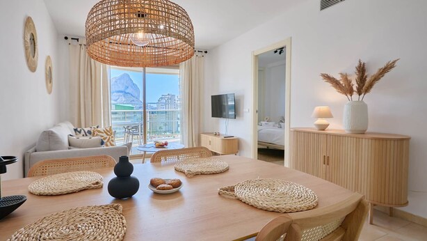 Una sala de estar moderna con una mesa de comedor de madera, individuales tejidos, una lámpara colgante grande de fibras naturales y vista a las montañas a través de la ventana.