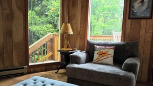 A cozy room with wooden walls and floors, a grey sofa, a matching ottoman, a small table with a lamp, and a painting on the wall. - New Kitchen! ‖ Hot Tub coming August 7th! ‖ 2 Mins to Slopes ‖ Paved Road Access (Beech Mountain)