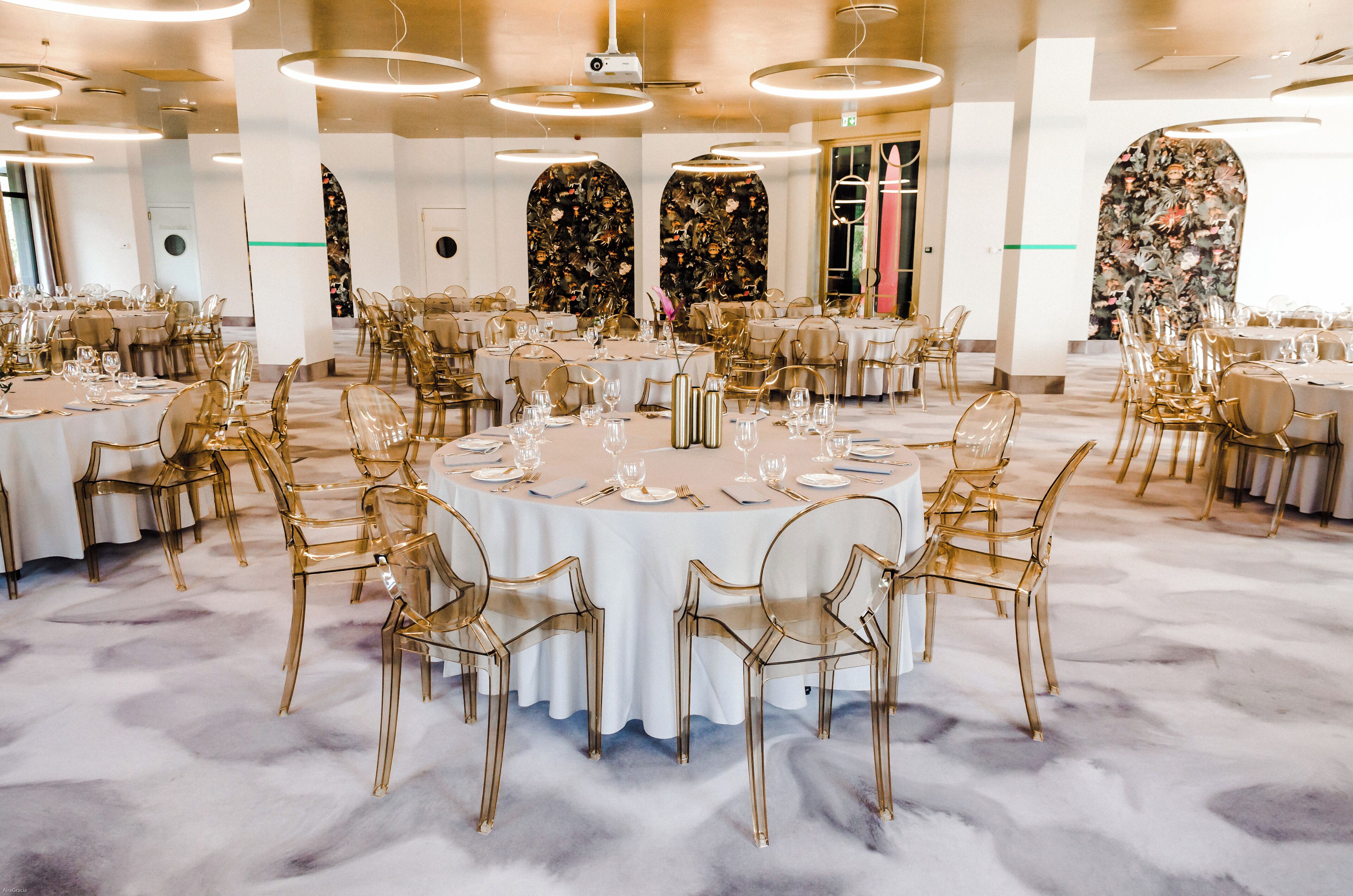 Een balzaal met ronde tafels, transparante stoelen en witte tafelkleden.