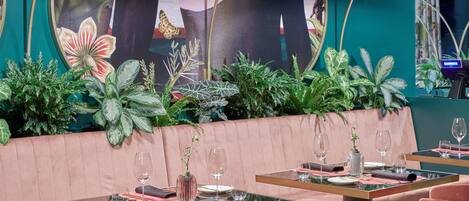 Un restaurante con una zona de asientos en una cabina rosa, mesas con sillas y un espejo grande con una pintura floral.