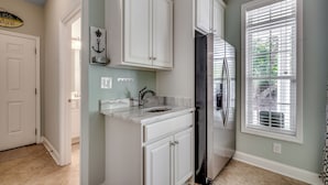 A kitchen with white cabinets, a stainless steel refrigerator, a marble countertop, and a window with blinds.