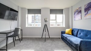 A modern living room with a grey carpet, a blue sofa with a yellow pillow, a grey chair, a tripod stand, and a flat-screen TV mounted on the wall. - Marygate by Deuce Hotels Ltd (Wakefield)