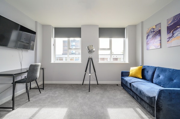 Una sala de estar moderna con alfombra gris, un sofá azul con un cojín amarillo, una silla gris, un trípode y un televisor de pantalla plana montado en la pared.