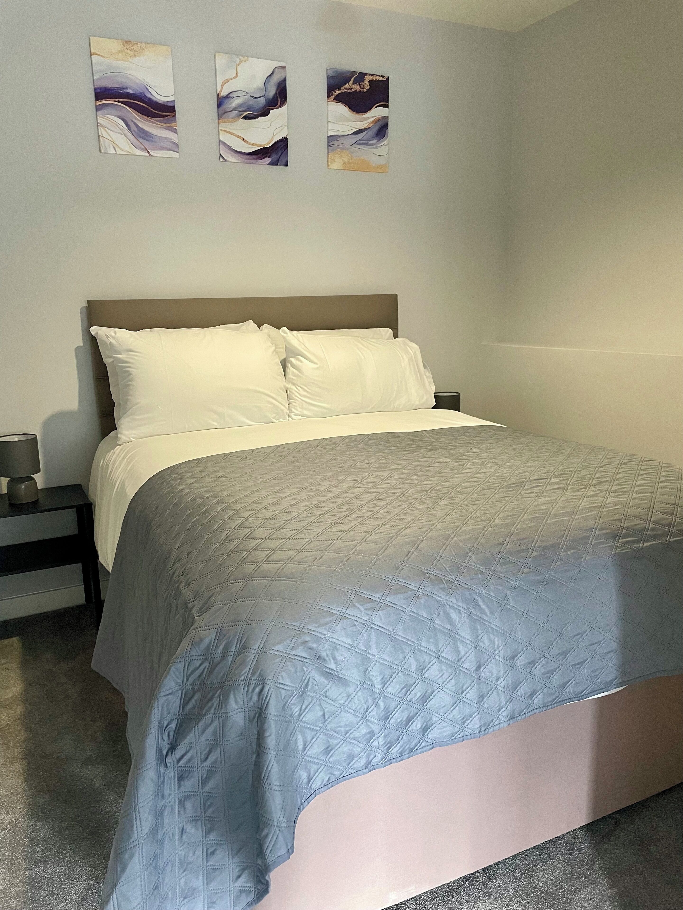 A neatly made bed with a quilted cover, a bedside table with a lamp, and three abstract paintings on the wall.