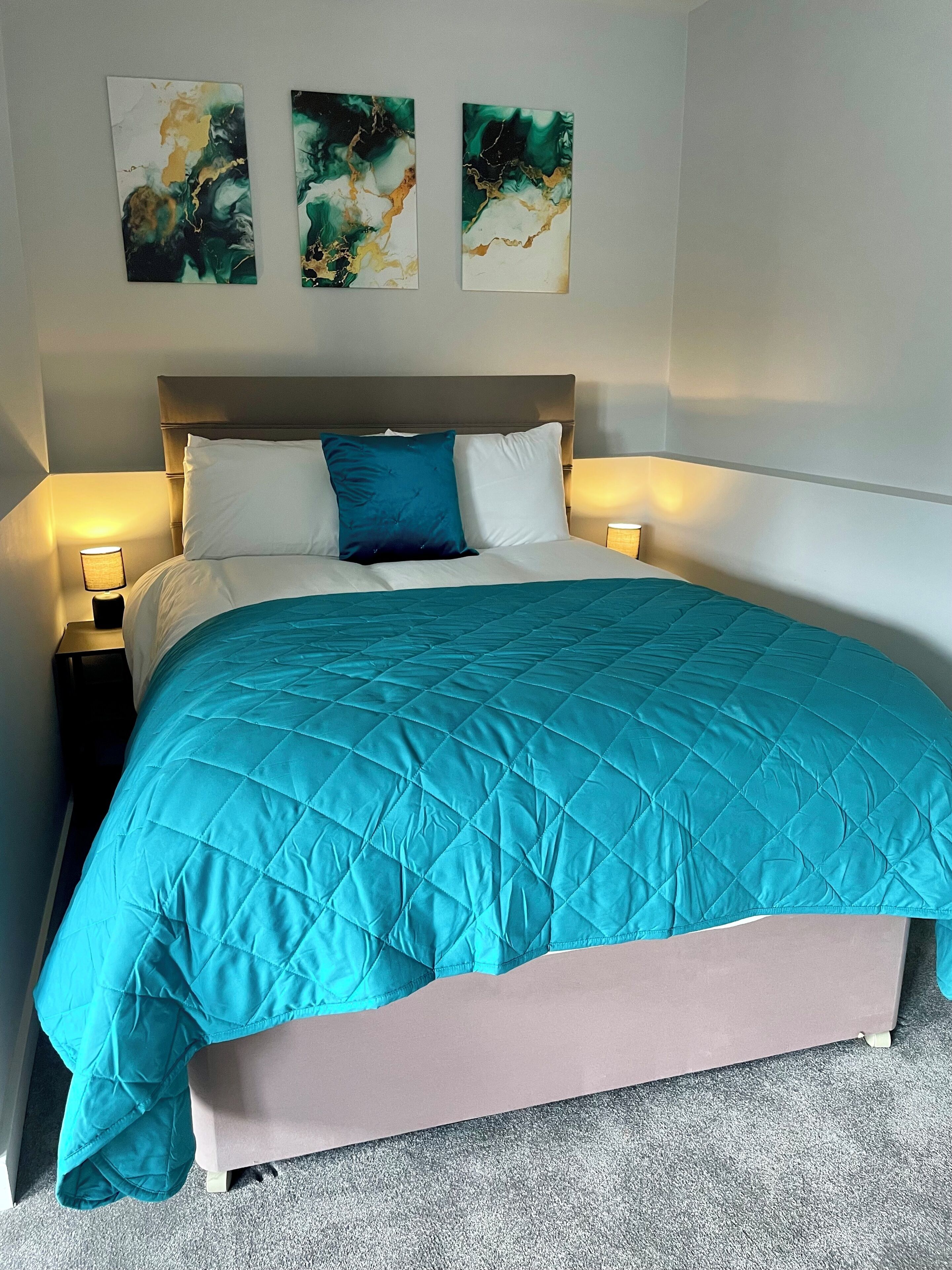 A neatly made bed with a teal quilt, two white pillows, and a blue accent pillow. There are three abstract paintings on the wall above the bed.