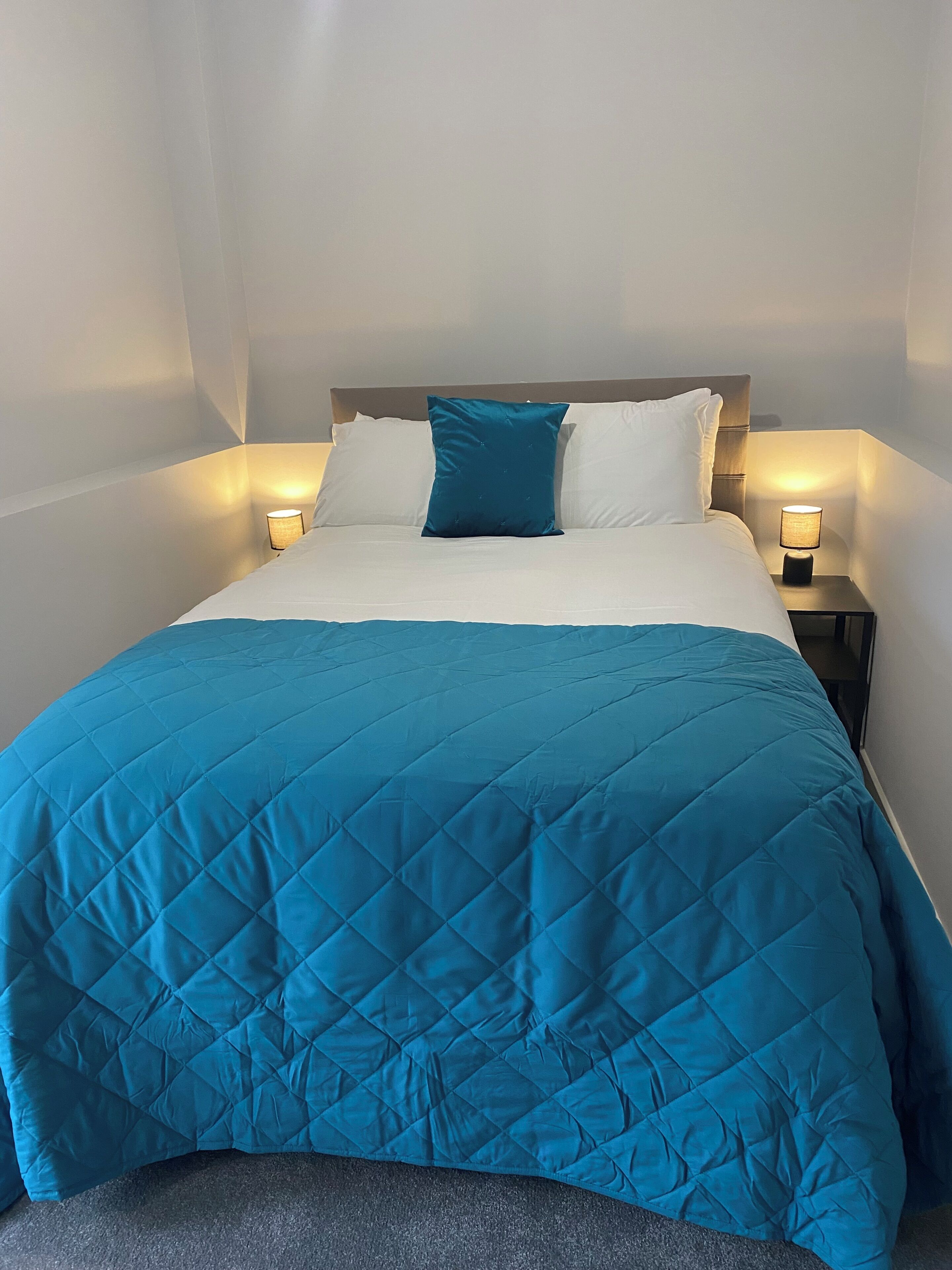 Una cama bien hecha con colcha azul, almohadas blancas y dos lámparas de noche.