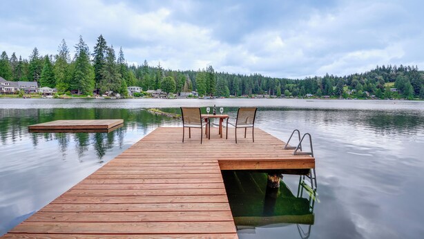 A wooden dock extending into calm water with a table and chairs, surrounded by dense forest and houses in the background. - Lakefront Snohomish Home ~ 13 Mi to Historic Dtwn (Snohomish)