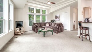 A spacious living room with a large window, a ceiling fan, a television on a stand, a green coffee table, a brown sofa, and a kitchen area with wooden cabinets. - Lakefront Snohomish Home ~ 13 Mi to Historic Dtwn (Snohomish)