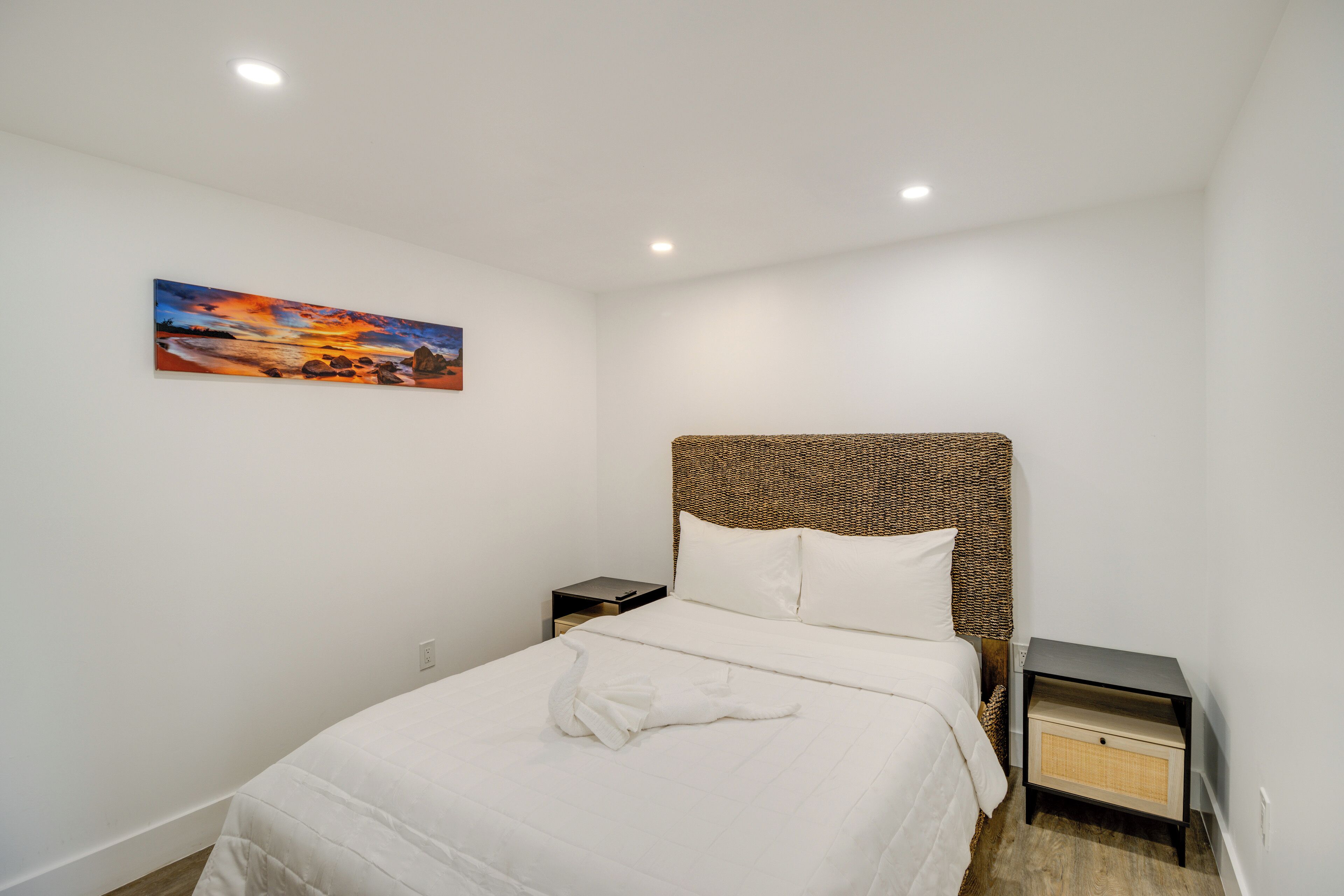 Un lit bien fait, avec une literie blanche, une tête de lit ornée d’un motif tissé, une table de chevet avec un tiroir et un tableau représentant un coucher de soleil accroché au mur.
