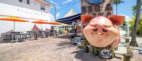 Un espacio al aire libre con pavimento de ladrillo, mesas y sillas, una gran escultura de un cerdo y un letrero que dice 'BBQ & Pizza'.