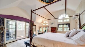 Ein Bett mit Baldachin, mit weißer Bettdecke, eine Holzbank und ein kleiner Tisch mit Stuhl in einem Zimmer mit großen Fenstern und Blick ins Freie.