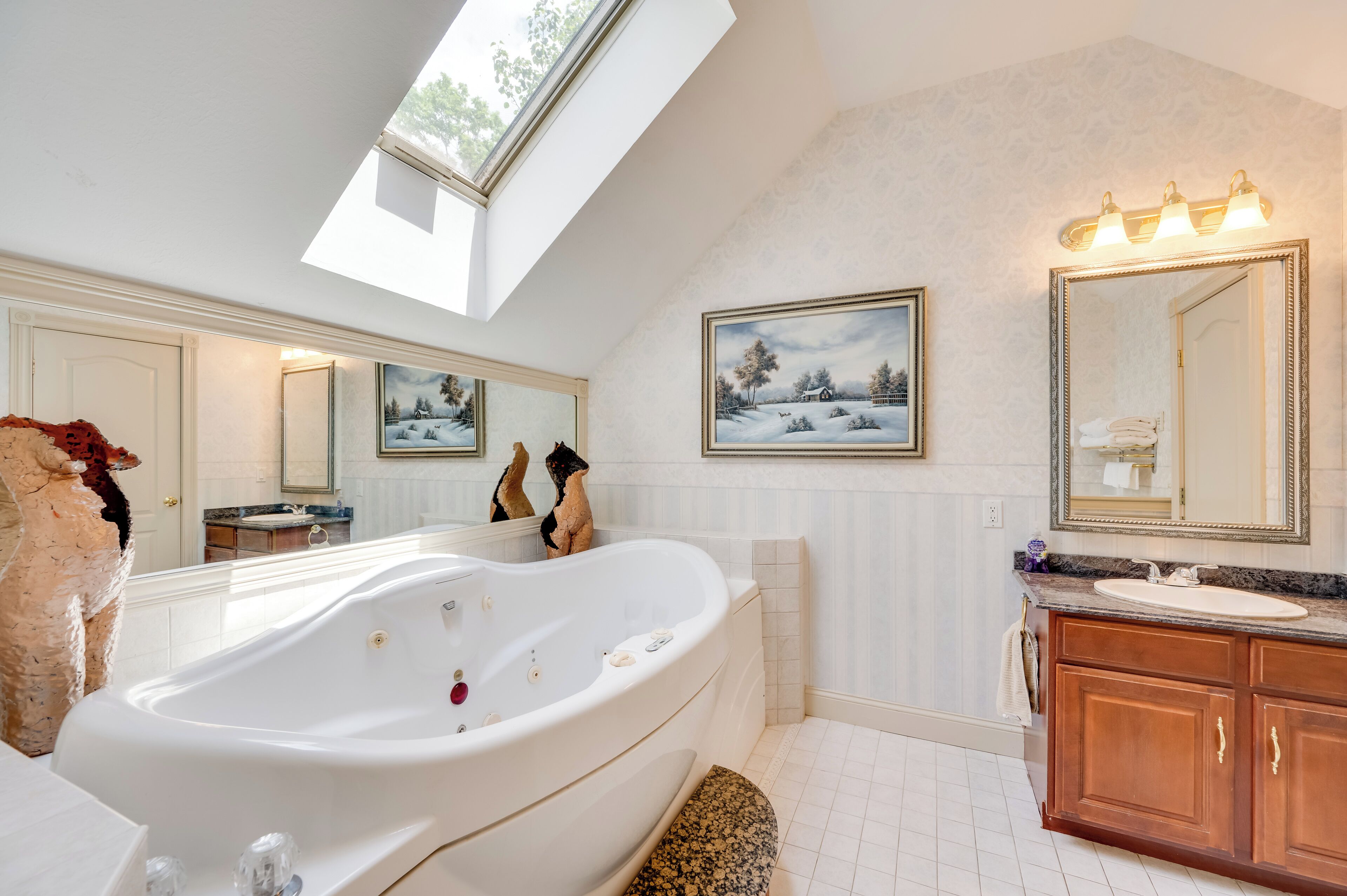 Une salle de bain spacieuse avec une grande baignoire sur pieds, un meuble-lavabo et un grand miroir. Une lucarne se trouve au-dessus de la baignoire et un tableau orne le mur.
