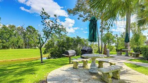 Un patio exterior bien cuidado con mesas y bancos de piedra, rodeado de exuberante vegetación y un cielo azul despejado.