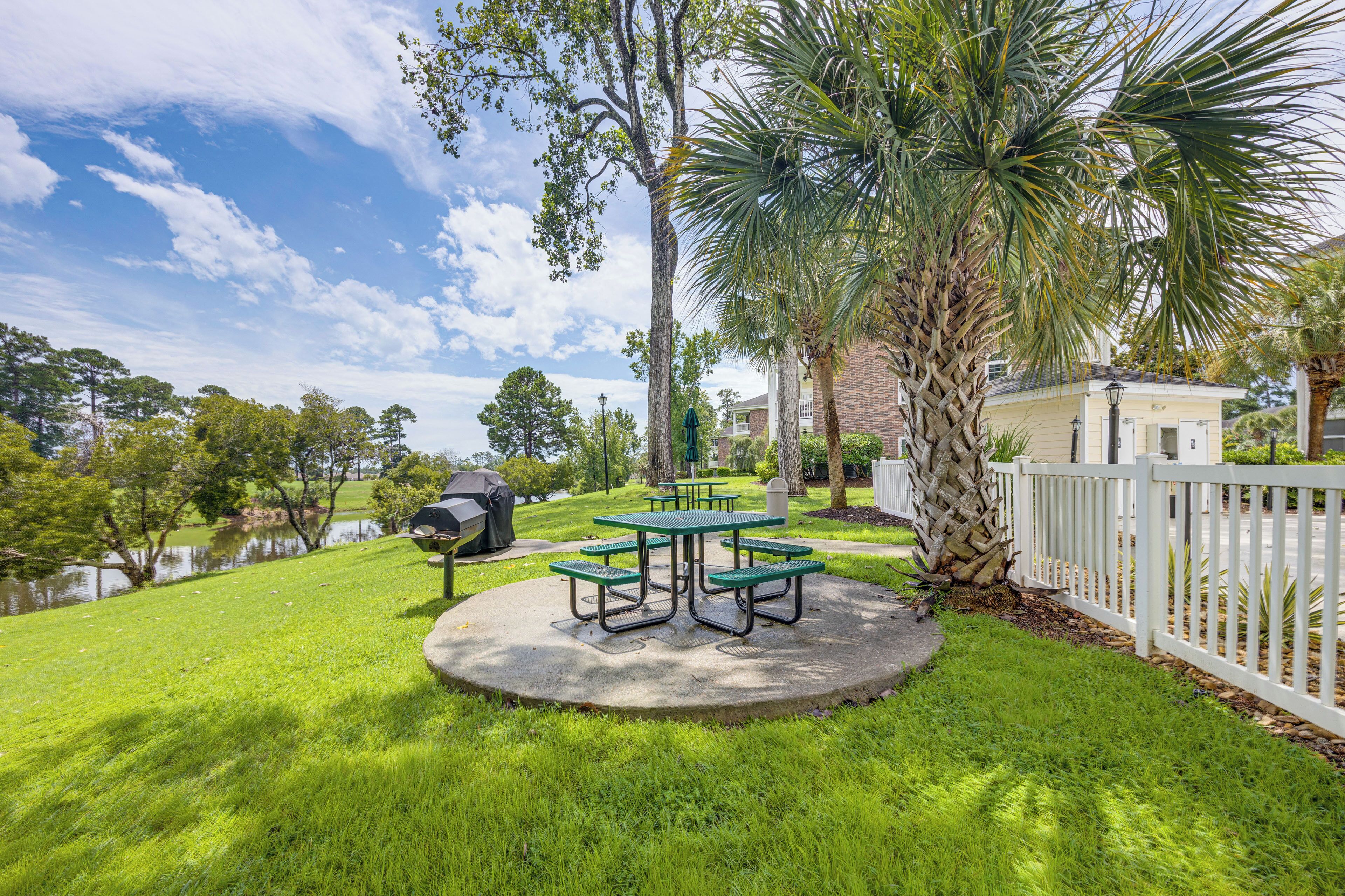A grassy area with picnic tables and a barbecue grill, surrounded by trees and a white fence.