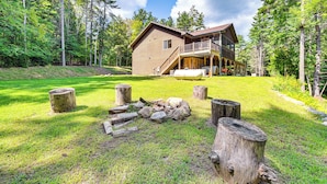 A cabin with a deck, surrounded by a forest and a grassy yard with a fire pit. - Tree Tops Retreat ~ 19 Mi to Lake George! (Chestertown)