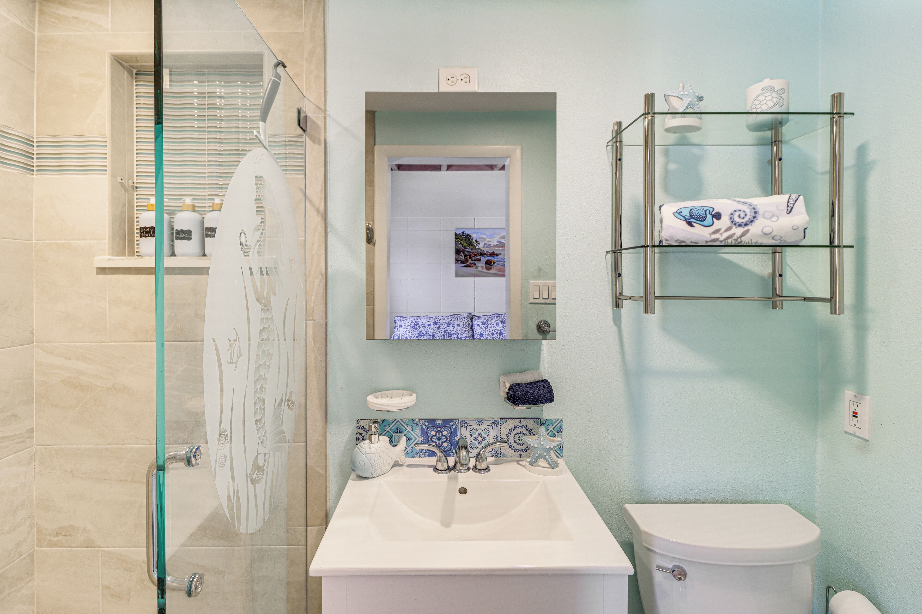 A bathroom with a glass shower enclosure, a white sink, and a toilet. There is a glass shelf with towels and toiletries.
