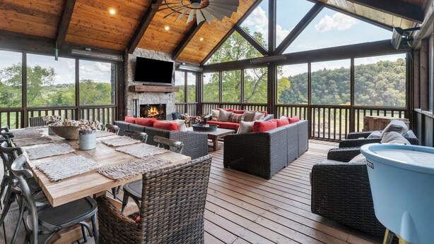 A covered outdoor area with a wooden deck, wicker furniture, a fireplace, and a large window overlooking a forested area. - Above Nashville Rustic Retreat Farm I 200 Acre Farm | Pool&Spa I Services (Charlotte)