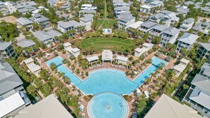 An aerial view of a resort with a central swimming pool surrounded by villas, palm trees, and a well-maintained lawn.