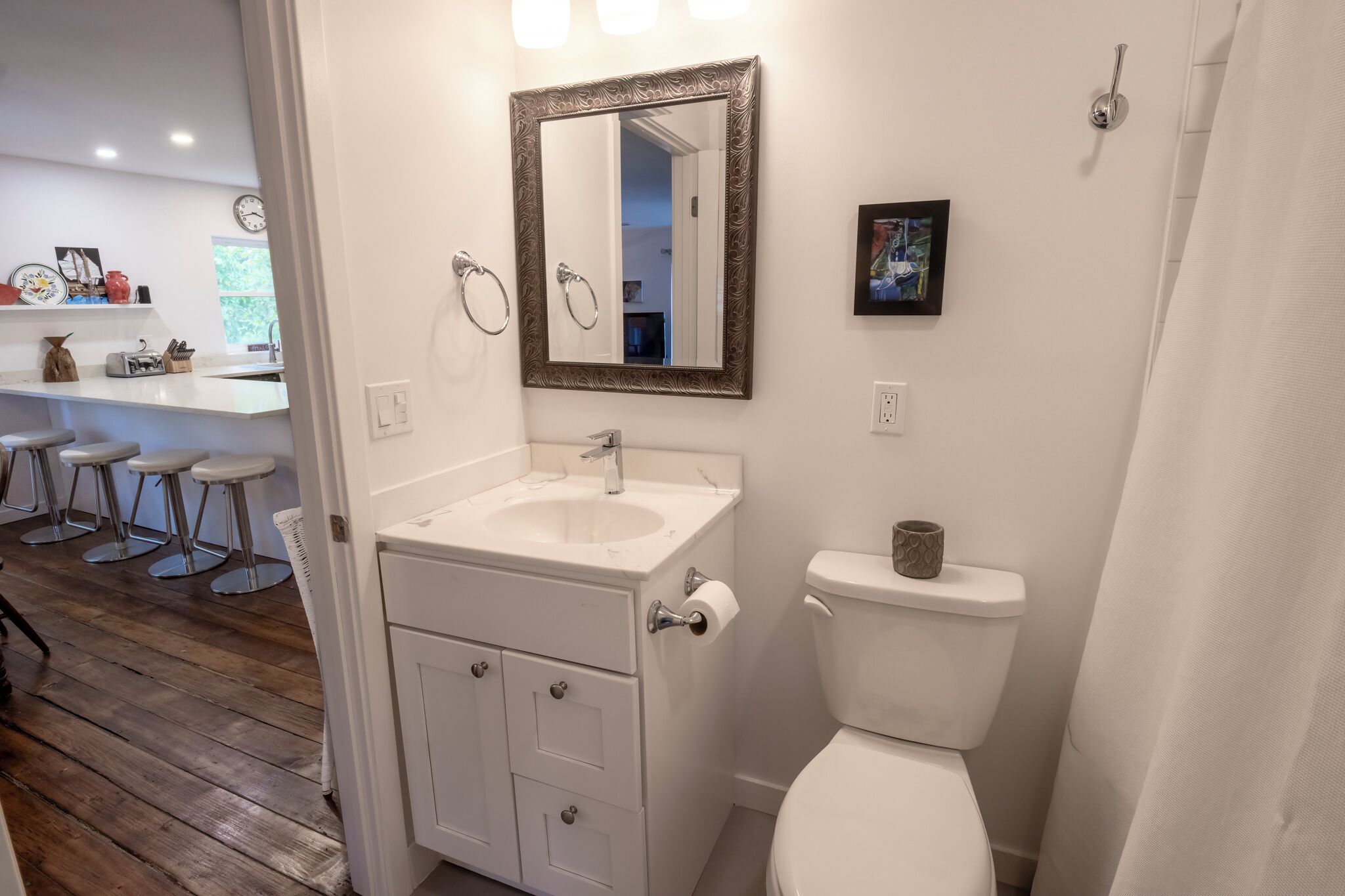 Un baño con lavamanos, inodoro y espejo blancos. Se ve un ventanal con vistas al exterior, lleno de vegetación.