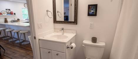 Un baño con lavamanos, inodoro y espejo blancos. Se ve un ventanal con vistas al exterior, lleno de vegetación.