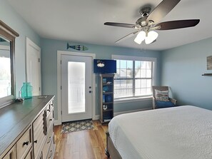 A bedroom with a bed, a ceiling fan, a window with blinds, a bookshelf, and a door with frosted glass.