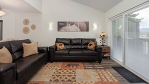 A living room with a black leather sofa, a patterned rug, and a large window with blinds.