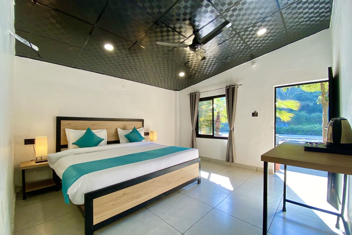A modern hotel room with a large bed, a ceiling fan, a window with curtains, and a small table with a kettle.