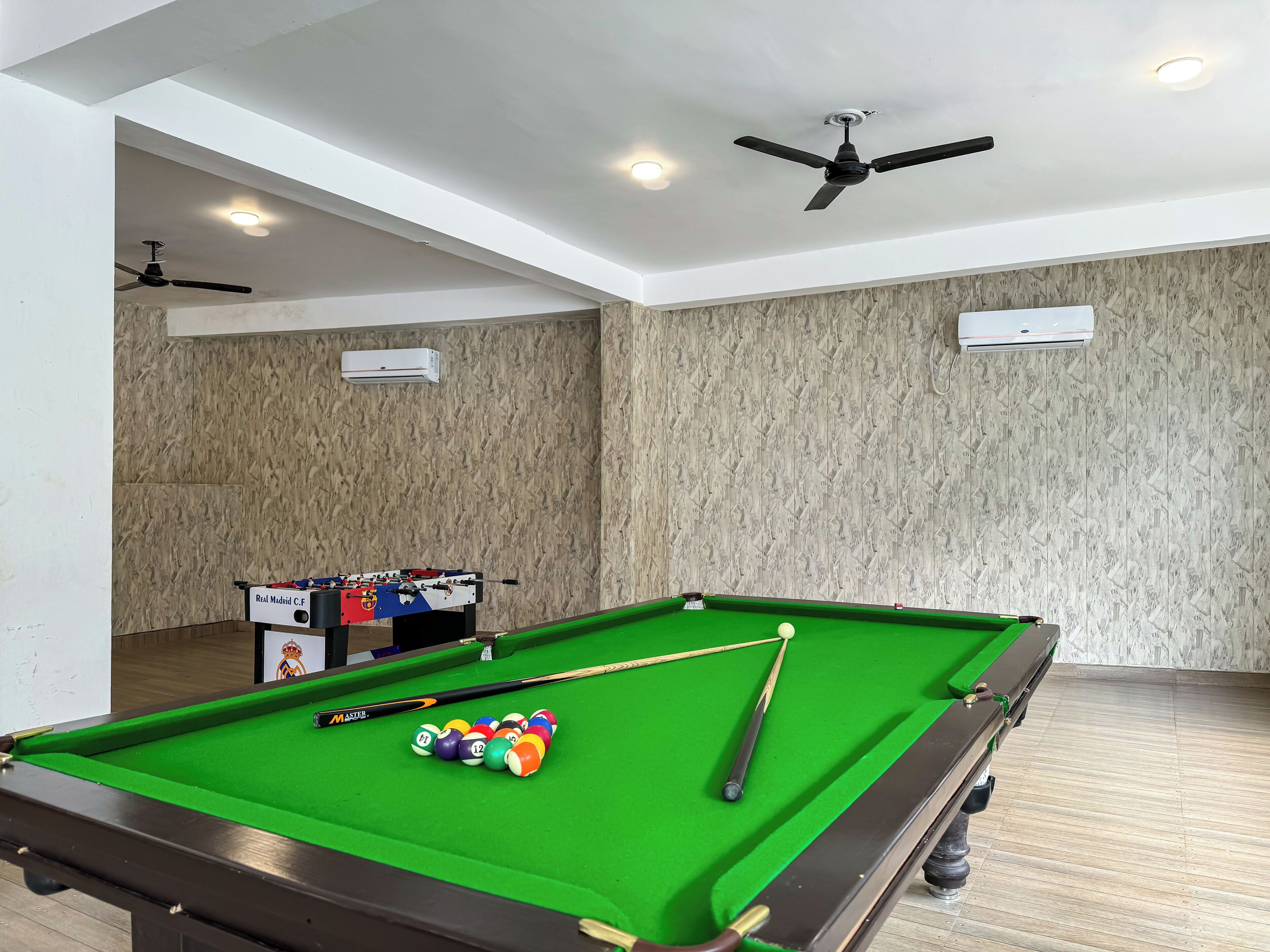 Mesa de billar con bolas, mesa de fútbol de mesa y ventiladores de techo en una habitación con paredes de madera e iluminación empotrada.