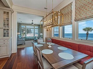 Un vaste espace dédié à la salle à manger, avec une table et des bancs en bois, de grandes fenêtres offrant une vue sur l’océan et un lustre décoratif.