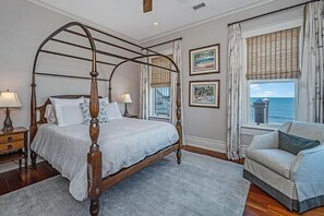 Un lit à baldaquin avec un cadre en bois, un tapis gris, un fauteuil gris et une fenêtre donnant sur la mer.