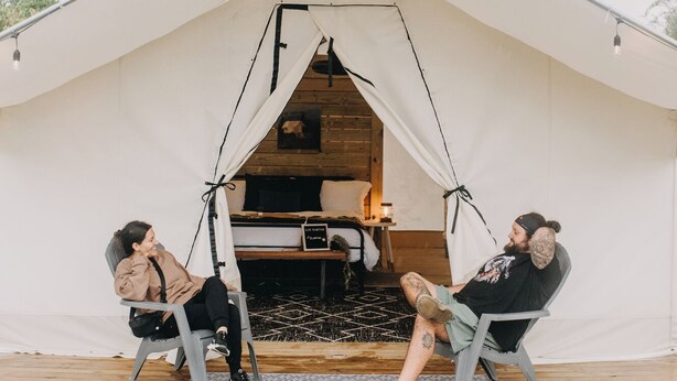 A tent with a bed inside, two people sitting on chairs outside, and a grassy area in front.