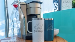 Une table en bois, une bouteille en verre, une tasse blanche et une tasse bleue ornée d’un logo représentant un ours.