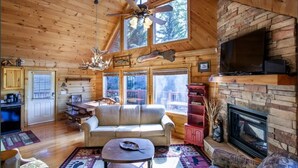 A log cabin interior with a fireplace, a television, a ceiling fan, and a dining area.