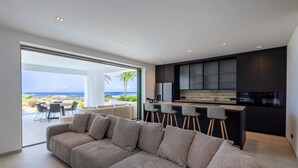 Una sala moderna con un amplio sofá modular, una cocina con armarios oscuros y una barra con taburetes.