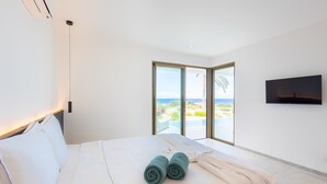 Un dormitorio moderno con una cama grande, un televisor montado en la pared y vista al océano a través de un ventanal corredera.