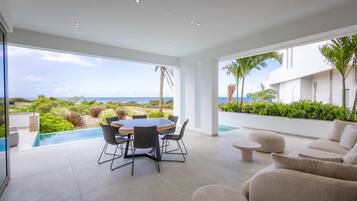 Un área de comedor exterior moderna con piscina, con vistas a un paisaje exuberante y el mar.