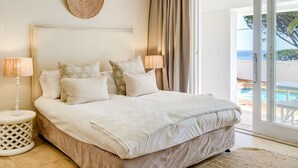 A bedroom with a large bed, two bedside lamps, a woven wall decoration, and a view of a pool through a sliding glass door. - Classic Charm Meets Modern Comfort at Atlantic Pearl (Cape Town)