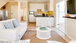 A modern kitchen with white cabinets, a central island with stools, and a dining area with a white table and chairs. - Classic Charm Meets Modern Comfort at Atlantic Pearl (Cape Town)