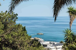 A coastal view with a rocky shoreline, a clear blue sea, and a building with solar panels. - Classic Charm Meets Modern Comfort at Atlantic Pearl (Cape Town)