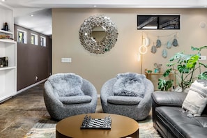Un salon avec deux fauteuils gris, une table basse sur laquelle se trouve un jeu d’échecs, un miroir et une plante.