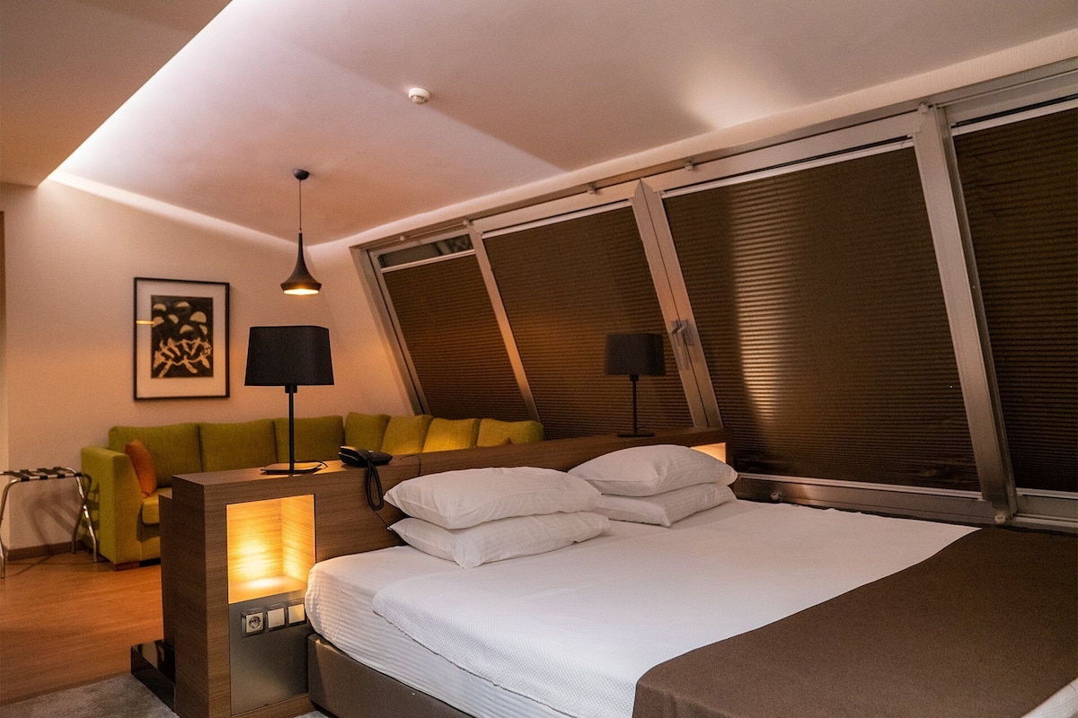 A modern bedroom with a large bed, a desk with a lamp, a sofa, and a window with blinds.
