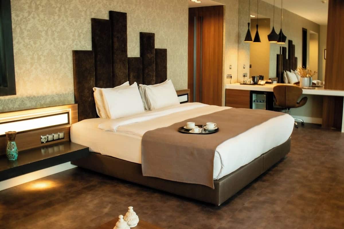 A modern hotel room with a large bed, bedside table, and a desk area with a chair.