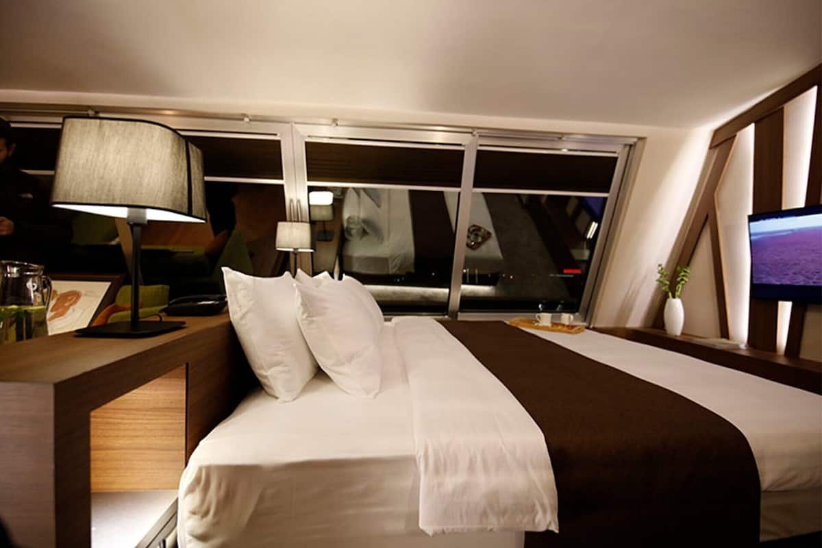 A hotel room with a bed, a nightstand, a lamp, a TV, and a window.